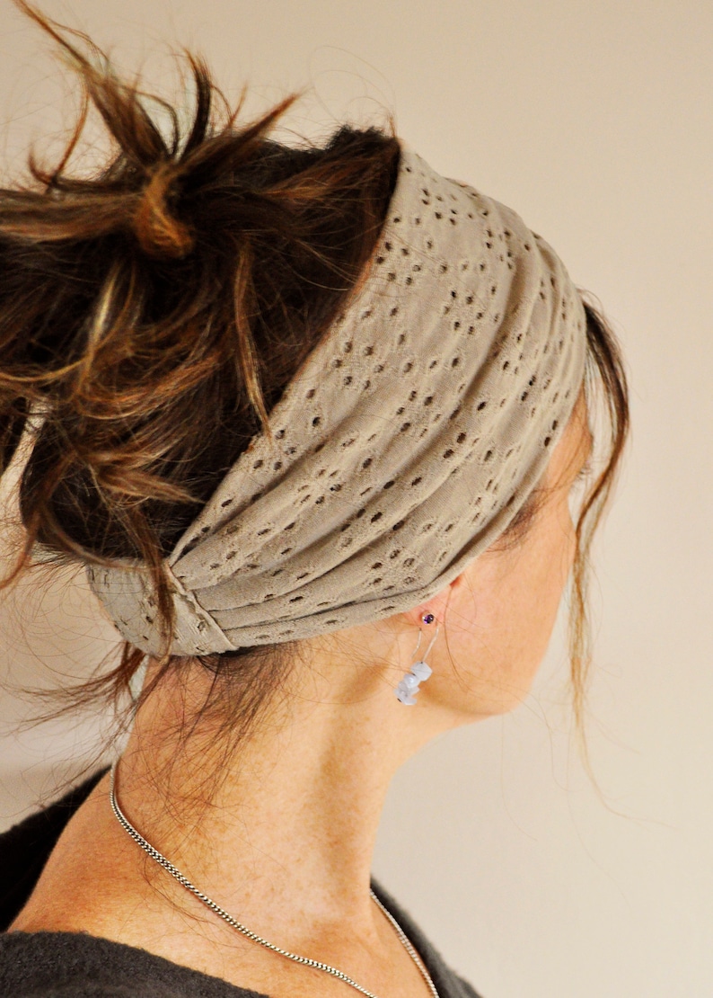 May include: A wide, taupe-colored headband featuring a repeating eyelet lace pattern with small, circular cutouts. The fabric is gathered and draped, creating a soft, textured appearance that wraps around the head in a simple, functional design.