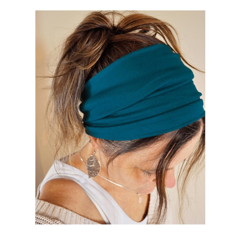 May include: A teal-coloured wide headband made of soft fabric. The headband is wrapped around the head, covering the forehead and ears. The image shows a close-up of the headband, with the person's hair pulled back in a bun. The headband is a solid colour and appears to be comfortable.