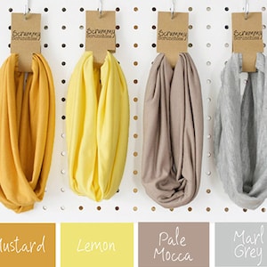 May include: Four infinity scarves in mustard, lemon, pale mocca, and marl grey. Each scarf is displayed on a pegboard with a brown tag that reads "Scrummy Scrunchies". The colour names are displayed below the scarves.