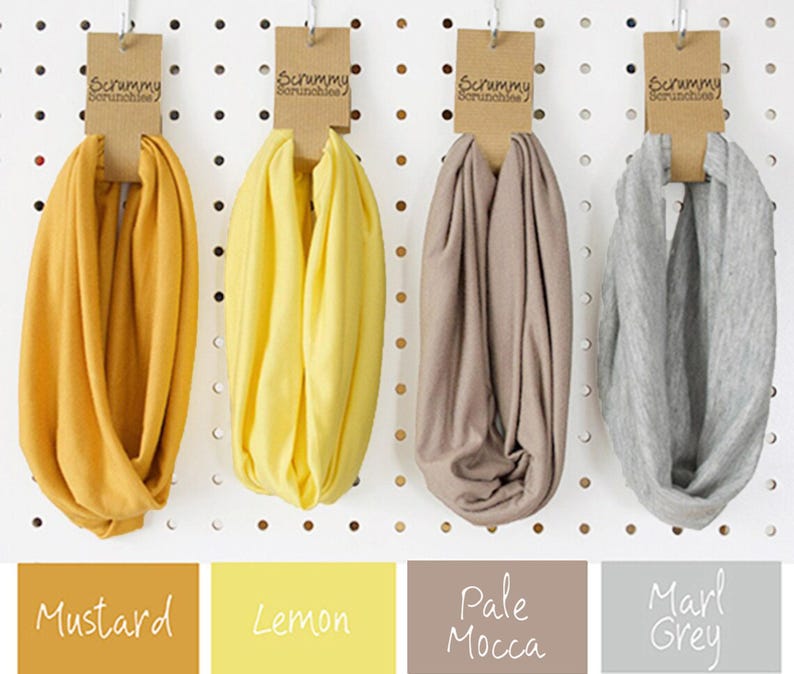 May include: Four infinity scarves in mustard, lemon, pale mocca, and marl grey. Each scarf is displayed on a pegboard with a brown tag that reads "Scrummy Scrunchies". The colour names are displayed below the scarves.