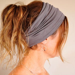 May include: A gray fabric headband worn around the head. The headband is wide and gathered, with a soft, draped appearance. The person is also wearing a silver necklace and earrings.