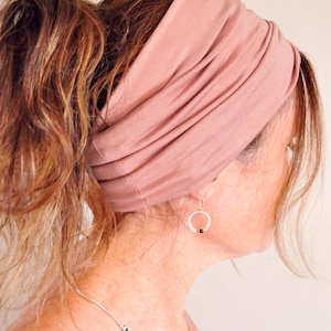May include: A wide headband in a dusty rose hue is worn. The headband is made of a soft, stretchy material. A silver necklace and earrings with a small red stone are also visible.