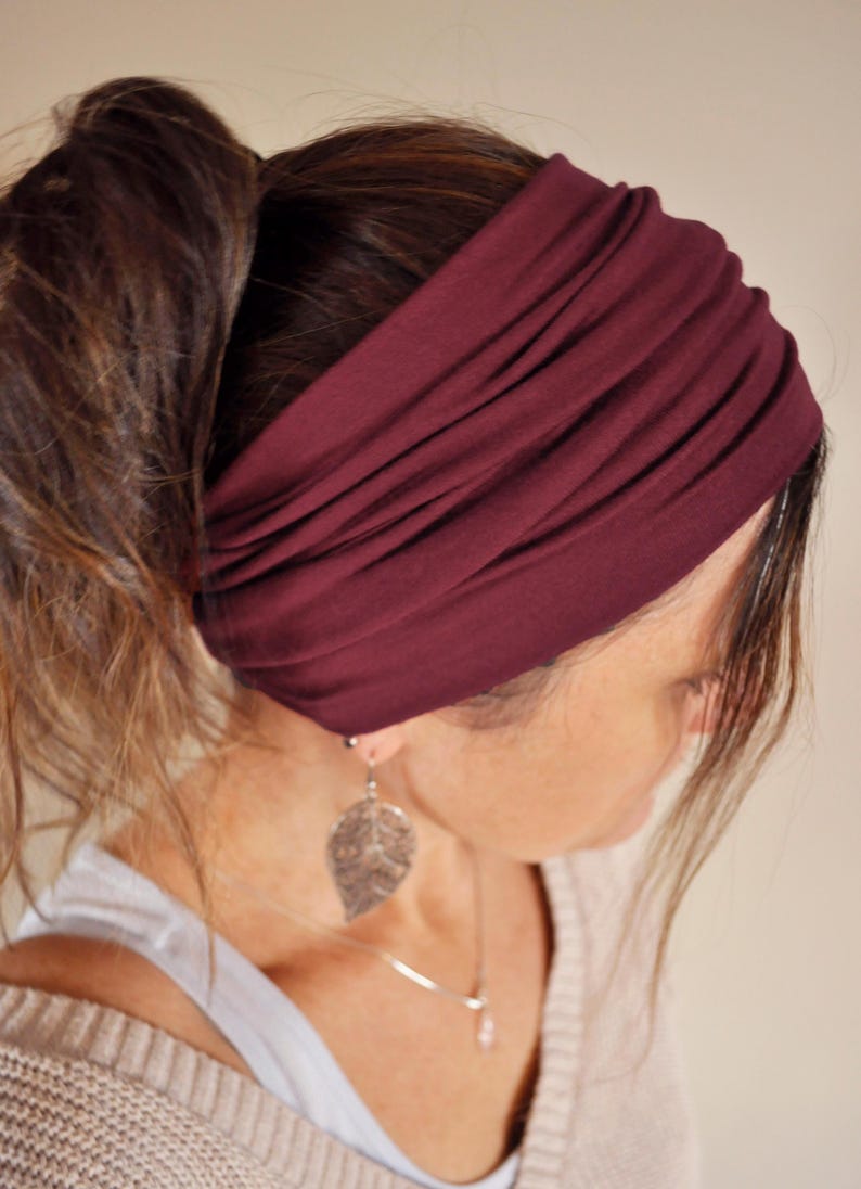 May include: A burgundy-colored wide headband is worn across the forehead. The fabric is gathered, creating a textured appearance. The headband is paired with a silver leaf-shaped earring and a silver necklace. The hair is pulled back in a ponytail.