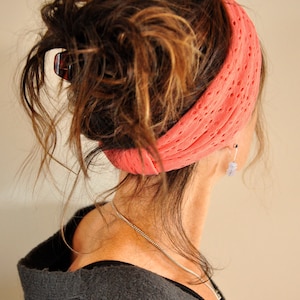 May include: A woman with long, brown hair wearing a coral-colored knitted headband. Her hair is pulled back in a messy bun. She is wearing a gray sweater.