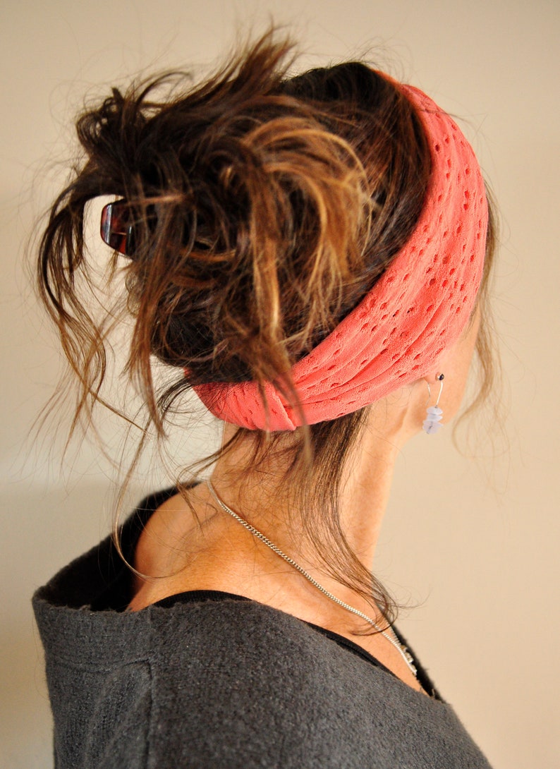 May include: A woman with long, brown hair wearing a coral-colored knitted headband. Her hair is pulled back in a messy bun. She is wearing a gray sweater.