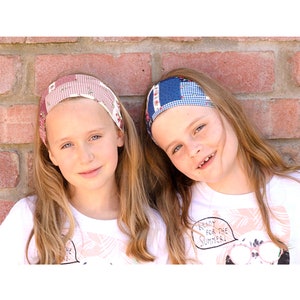 以下が含まれることがあります： 白いTシャツとカラフルなヘッドバンドをつけた、長いブロンドの髪の女の子2人。左側の女の子はピンクと白のヘッドバンドをつけています。右側の女の子は青と白のヘッドバンドをつけています。右側の女の子のシャツには「夏に備えよう！」という文字が印刷されています。