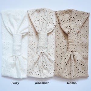 May include: Three wide headbands featuring an eyelet lace pattern with a central knotted detail. The headbands are displayed in three distinct colors labeled as Ivory, Alabaster, and Mocha, arranged from left to right against a plain white background.