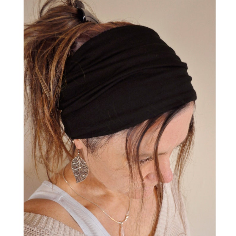 May include: A black wide headband made of soft fabric. The headband is worn around the forehead and covers the top of the head. The hair is pulled back and up, secured with a black clip. A silver leaf earring is visible.