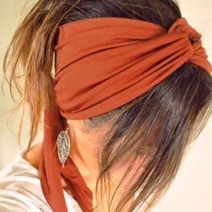 May include: A wide, rust-colored fabric headband featuring a twisted knot detail at the front. The headband is made of a smooth, solid-colored material that wraps around the head, with long, trailing fabric ends hanging down on one side.