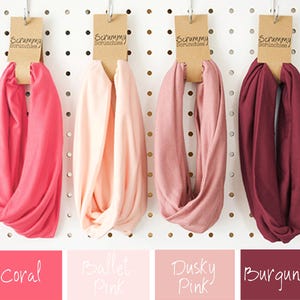 May include: Four fabric scrunchies in coral, ballet pink, dusky pink, and burgundy hang from a pegboard. Each scrunchie is attached to a brown tag with the text "Scrummy Scrunchies". The color names are displayed below each scrunchie.