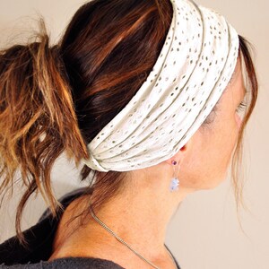 May include: A wide white headband featuring a delicate eyelet lace pattern with small circular cutouts. The fabric is gathered in horizontal pleats, creating a textured appearance that wraps around the head to secure the hair in place.