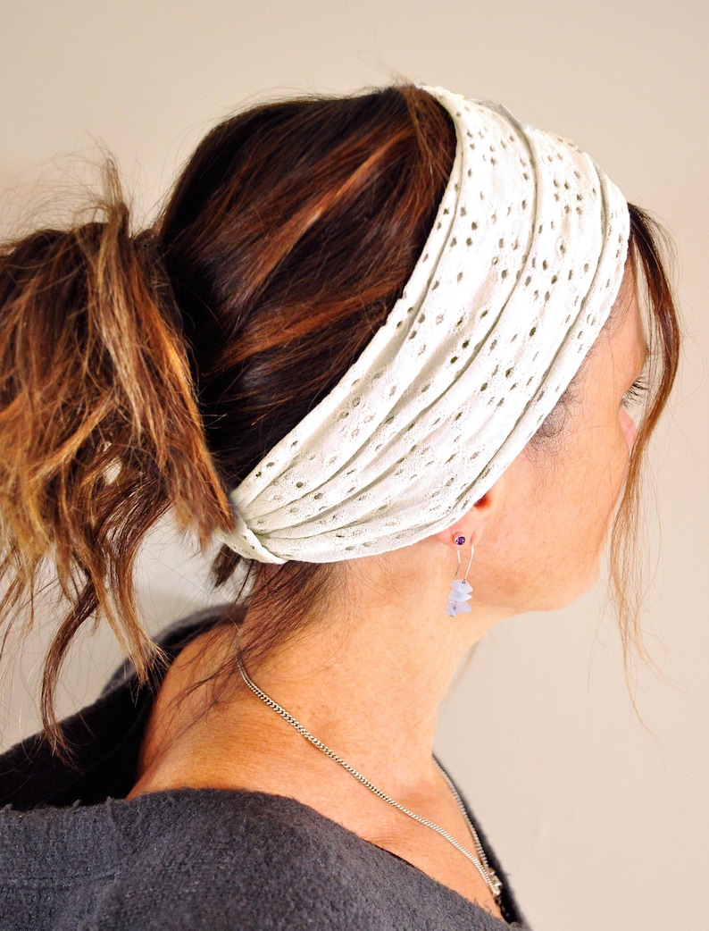 May include: A wide white headband featuring a delicate eyelet lace pattern with small circular cutouts. The fabric is gathered in horizontal pleats, creating a textured appearance that wraps around the head to secure the hair in place.