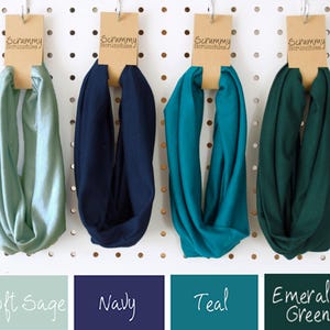May include: Four infinity scarves in Soft Sage, Navy, Teal, and Emerald Green hang from a pegboard. Each scarf is attached to a brown tag that reads "Scrummy Scrunchies". Below the scarves are color swatches with the names of the colors.