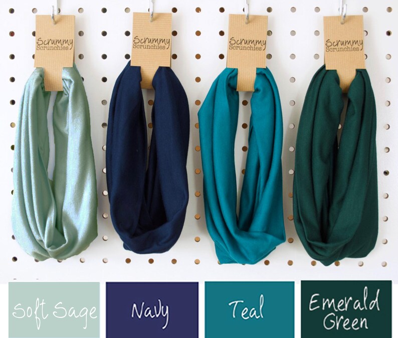 May include: Four infinity scarves in Soft Sage, Navy, Teal, and Emerald Green hang from a pegboard. Each scarf is attached to a brown tag that reads "Scrummy Scrunchies". Below the scarves are color swatches with the names of the colors.