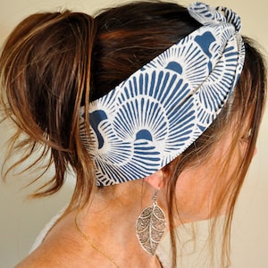 May include: A fabric headband with a blue and white pattern is worn around the head. The headband is knotted at the top. A silver leaf earring hangs from the ear. A gold necklace is also visible.