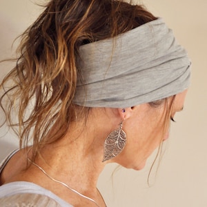 May include: A gray jersey knit headband worn around the head. The headband is wide and gathered, with the hair pulled back in a ponytail. The person is wearing a silver leaf-shaped earring and a silver necklace.