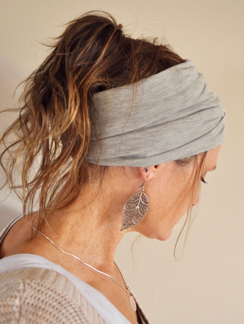May include: A gray jersey knit headband worn around the head. The headband is wide and gathered, with the hair pulled back in a ponytail. The person is wearing a silver leaf-shaped earring and a silver necklace.