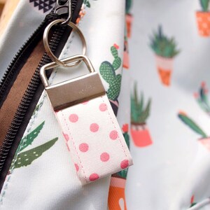 May include: A white backpack with a zipper closure and a pink polka dot keychain. The backpack has a cactus print.