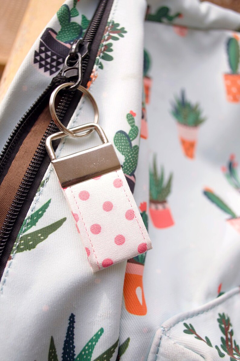 May include: A white backpack with a zipper closure and a pink polka dot keychain. The backpack has a cactus print.