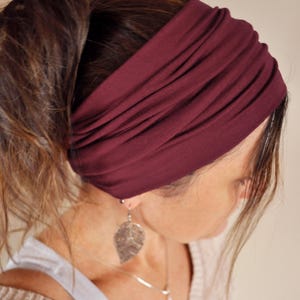 May include: A wide, burgundy-coloured headband made of soft fabric. The headband is worn across the forehead. The hair is pulled back into a ponytail. The person is wearing a silver leaf-shaped earring. The headband is approximately 10 cm wide.