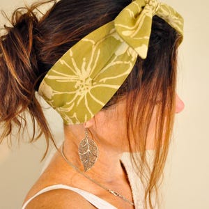 May include: A woman wearing a green and white floral print headband. The headband is tied in a bow on top of the head. She is also wearing a silver leaf-shaped earring and a silver necklace.