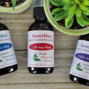 May include: Three brown glass aromatherapy spray bottles with black caps and white labels. The labels read "ScentMist" and feature phrases like "Help me Sleep," "Lift my Mood," and "Calm my Mind." The bottles are near small potted plants.