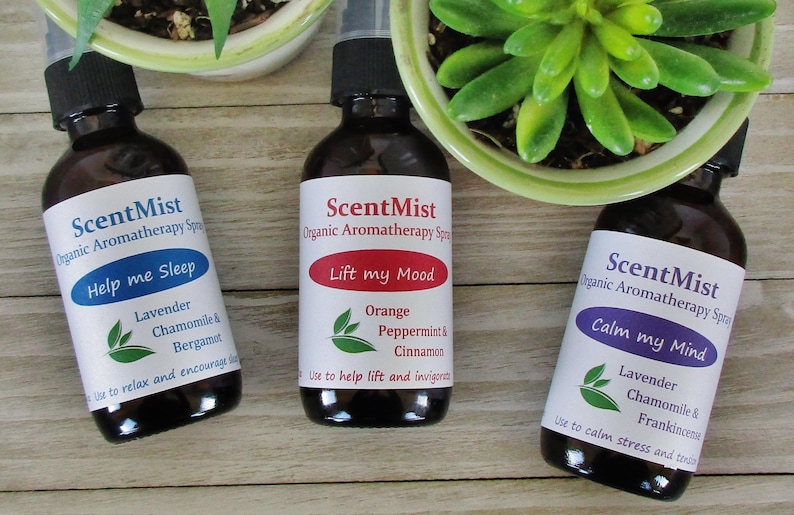 May include: Three brown glass aromatherapy spray bottles with black caps and white labels. The labels read "ScentMist" and feature phrases like "Help me Sleep," "Lift my Mood," and "Calm my Mind." The bottles are near small potted plants.
