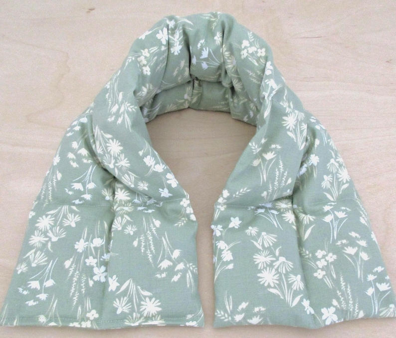 May include: A sage green neck wrap with a white floral pattern. The wrap is rectangular with a curved top and is designed to be worn around the neck. The fabric appears to be soft and filled with a weighted material.