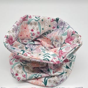 May include: A white fabric neck warmer with a floral pattern featuring pink, purple, and green flowers. The fabric is soft and comfortable.