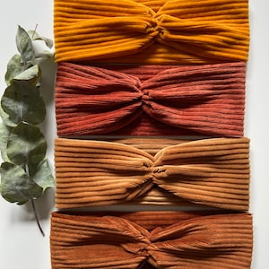 Pode incluir: Quatro faixas de cabelo de veludo cotelê em tons de ouro, ferrugem e castanho são dispostas em fila. Cada faixa apresenta um design de nó torcido. As faixas são feitas de tecido canelado e são exibidas em uma superfície branca.