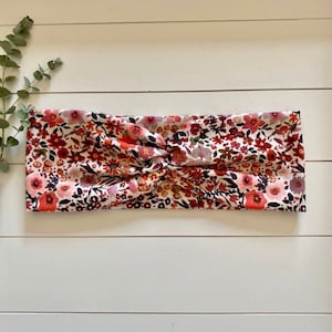 Könnte beinhalten: Ein weißes Stirnband mit einem Blumenmuster in Rot-, Rosa- und Brauntönen. Das Stirnband ist aus weichem, dehnbarem Stoff gefertigt und so konzipiert, dass es bequem zu tragen ist.