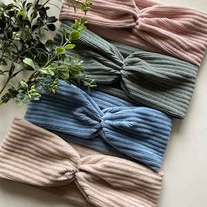 May include: Four fabric headbands in different colors: pink, green, blue, and beige. The headbands are made of a soft, ribbed material and have a knotted design.