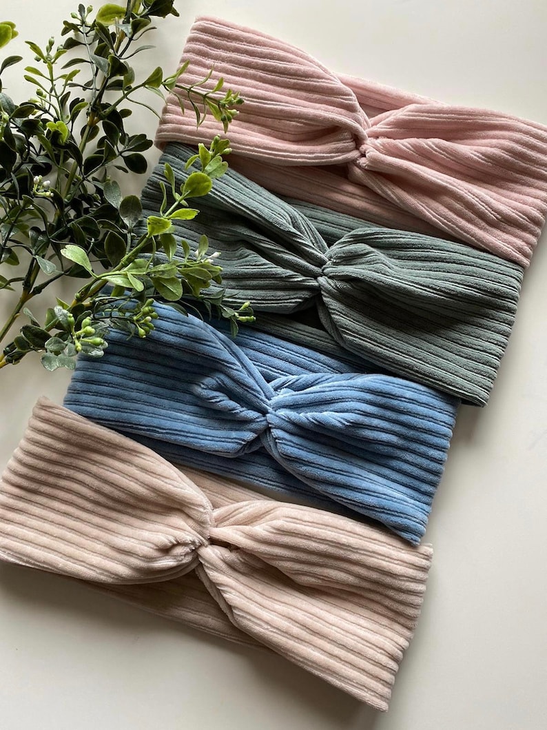 May include: Four fabric headbands in different colors: pink, green, blue, and beige. The headbands are made of a soft, ribbed material and have a knotted design.
