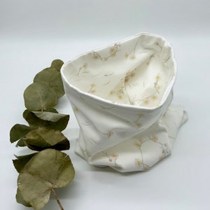 Peut inclure: Un snood en tissu blanc avec un motif floral. Le snood est plié, révélant un intérieur floral contrasté. Des feuilles d'eucalyptus séchées sont sur le côté gauche. Le snood est en tissu doux.