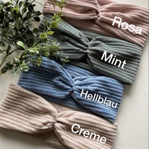 May include: Four fabric headbands in different colors: cream, light blue, mint green, and pink. Each headband has a knotted design and is labeled with its color name in German.