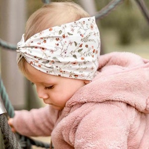 Pode incluir: Um bebê vestindo uma jaqueta rosa com capuz e uma faixa de cabeça com estampa floral branca. A faixa de cabeça está amarrada em um nó no topo da cabeça.