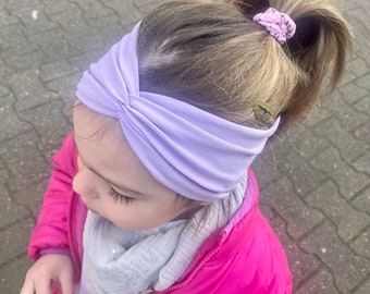May include: A young girl with blonde hair wearing a purple headband and a pink jacket. Her hair is pulled back in a ponytail with a pink hair tie.
