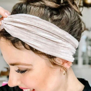 May include: A close-up of a woman wearing a light beige velvet headband. The headband is knotted at the top and has a subtle sheen. The woman has her hair pulled back in a messy bun.