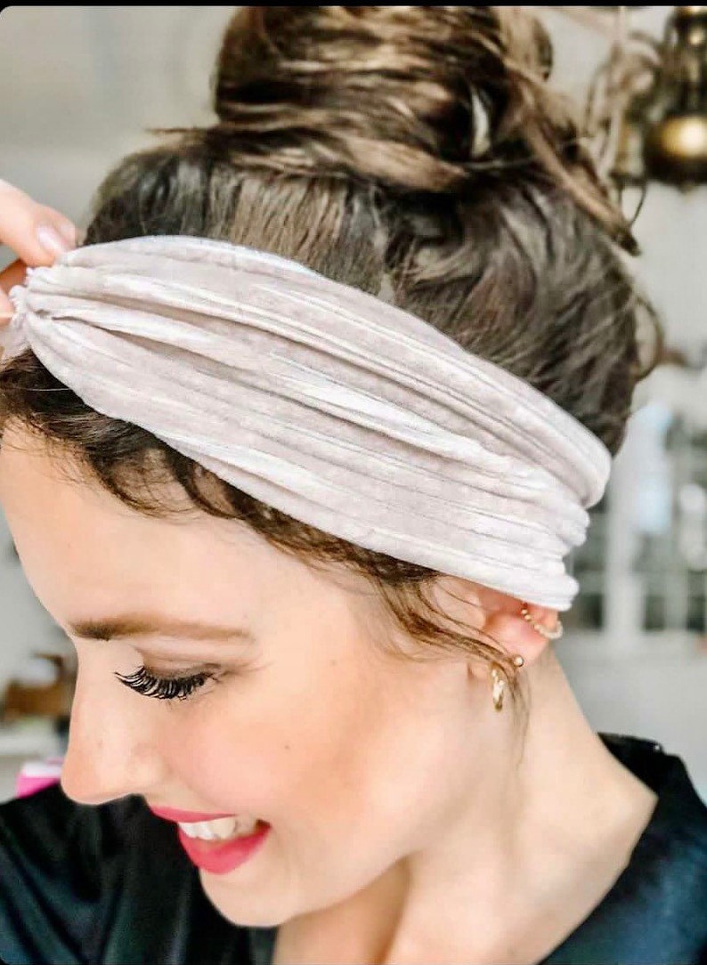 May include: A close-up of a woman wearing a light beige velvet headband. The headband is knotted at the top and has a subtle sheen. The woman has her hair pulled back in a messy bun.