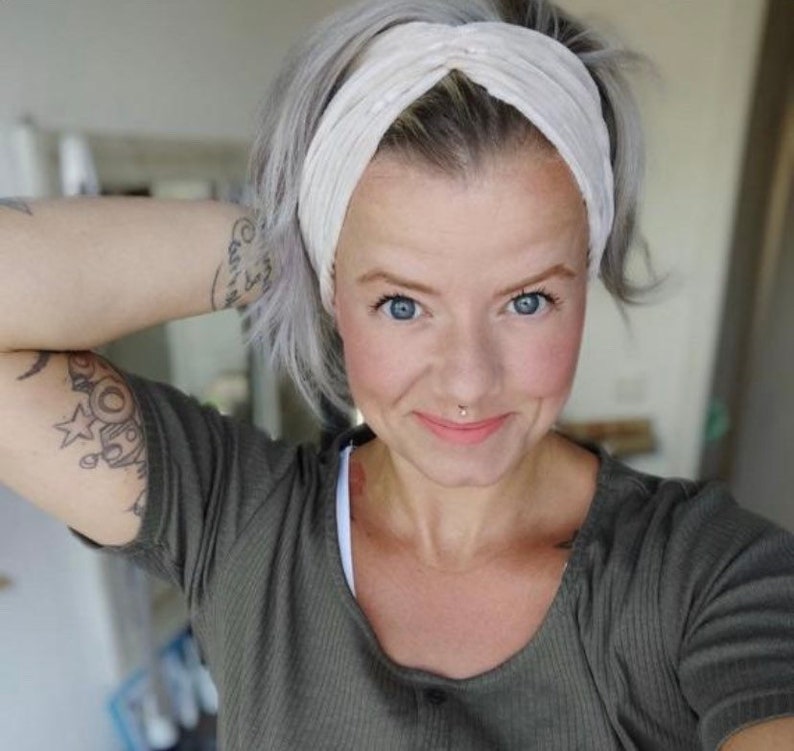May include: A woman with short, gray hair is wearing a white knotted headband. She is smiling and has a nose ring. She is wearing a green shirt.
