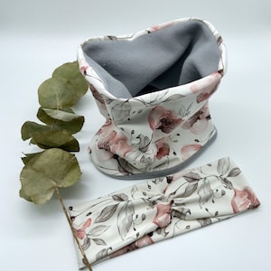 Op de afbeelding: Een witte hoofdband en nekwarmer set met een bloemenprint met roze en grijze bloemen. De hoofdband is in een geknoopte stijl en de nekwarmer is gevoerd met grijs fleece.