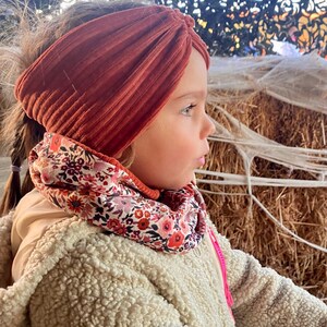 Op de afbeelding: Een jong meisje draagt een roestkleurig corduroy hoofdband en een nekwarmer met bloemenprint. Ze draagt een crèmekleurige fleecejas.