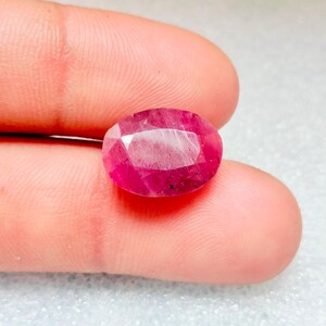 Puede incluir: Una piedra preciosa de rubí ovalada de color rosa vibrante con un corte facetado. La piedra preciosa se coloca sobre un fondo blanco.