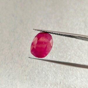 Puede incluir: Una gema ovalada de color rojo vibrante sostenida por unas pinzas. La gema tiene una apariencia ligeramente translúcida con un tono rojo intenso.