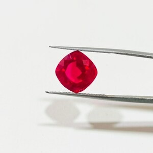 May include: A vibrant red gemstone with a cushion cut, held by tweezers against a white background.
