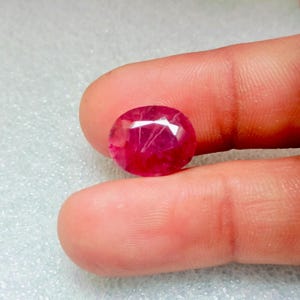 Puede incluir: Una gema de rubí rojo vibrante con forma ovalada facetada. La gema tiene una apariencia ligeramente translúcida con inclusiones visibles.