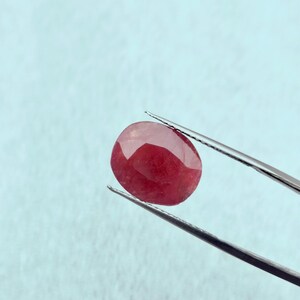 Puede incluir: Una piedra preciosa ovalada roja sostenida por unas pinzas sobre un fondo azul claro.