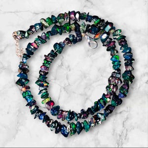 May include: A layered necklace featuring iridescent, irregularly shaped black opal stones. The stones display flashes of green, blue, and purple. The necklace has a silver-colored clasp and chain.