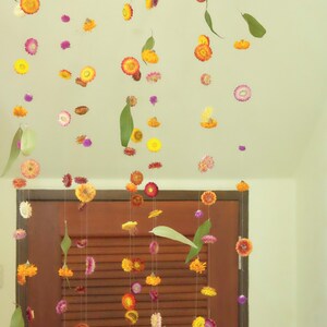 Peut inclure: Une guirlande de fleurs s&eacute;ch&eacute;es et de feuilles est suspendue devant une porte en bois. Les fleurs sont dans des tons de jaune, orange, rose et violet. Les feuilles sont vertes.