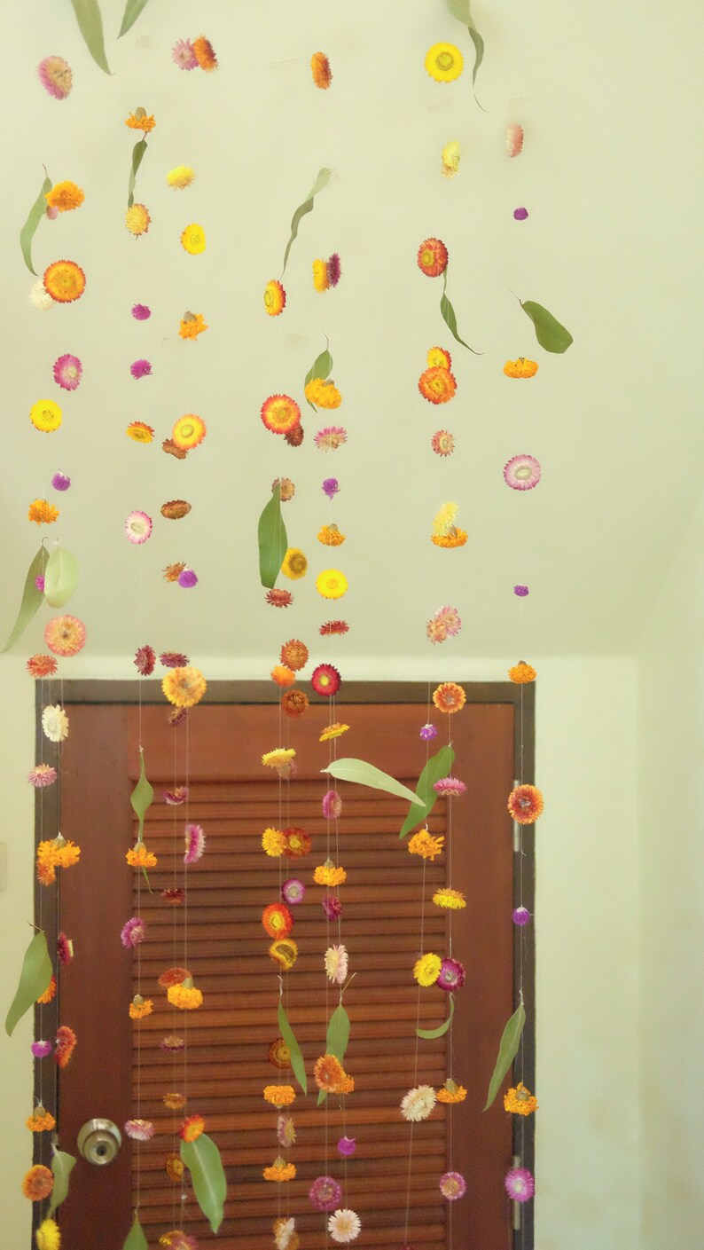 Peut inclure: Une guirlande de fleurs s&eacute;ch&eacute;es et de feuilles est suspendue devant une porte en bois. Les fleurs sont dans des tons de jaune, orange, rose et violet. Les feuilles sont vertes.